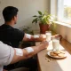 Two blood donors with white dressings on their arms sitting at a sunny window bench with glasses of milk and a plate of biscuits