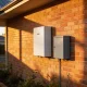 A white home battery unit and a grey box mounted on a brick wall in warm late-afternoon light, with rooftop solar panels at the top of the frame