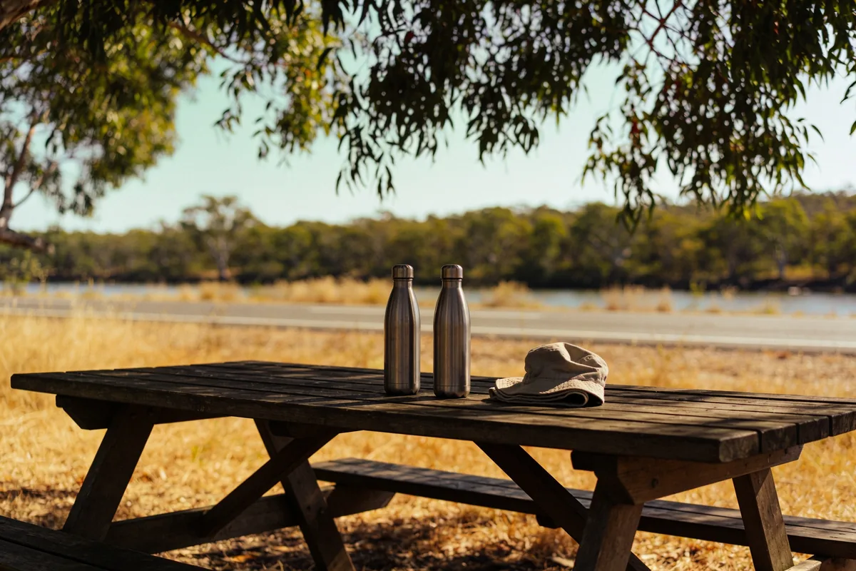 Two plain stainless steel water bottles and a folded cotton hat on a timber picnic table at a shaded roadside rest area, with dry grass and the highway behind