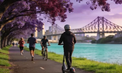 A commuter in a helmet riding an e-scooter along a riverside path past joggers and a cyclist, with jacaranda trees in bloom and the Story Bridge in the distance