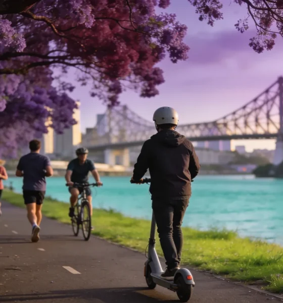 A commuter in a helmet riding an e-scooter along a riverside path past joggers and a cyclist, with jacaranda trees in bloom and the Story Bridge in the distance