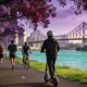A commuter in a helmet riding an e-scooter along a riverside path past joggers and a cyclist, with jacaranda trees in bloom and the Story Bridge in the distance