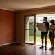A young couple standing in an empty, sunlit living room with timber floors, one holding a large sheet of paper, looking out through sliding doors to a grassed backyard