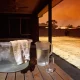 A shaded backyard patio on a hot afternoon with a tub of iced water and a wet face cloth, a jug of water and an empty glass on a timber table, beside a sun-bleached lawn under a hazy orange sky