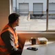 A worker in an orange hi-vis vest at a break room table with a coffee mug, a lunch box and a phone face down, looking out at the closed roller doors of a quiet loading dock