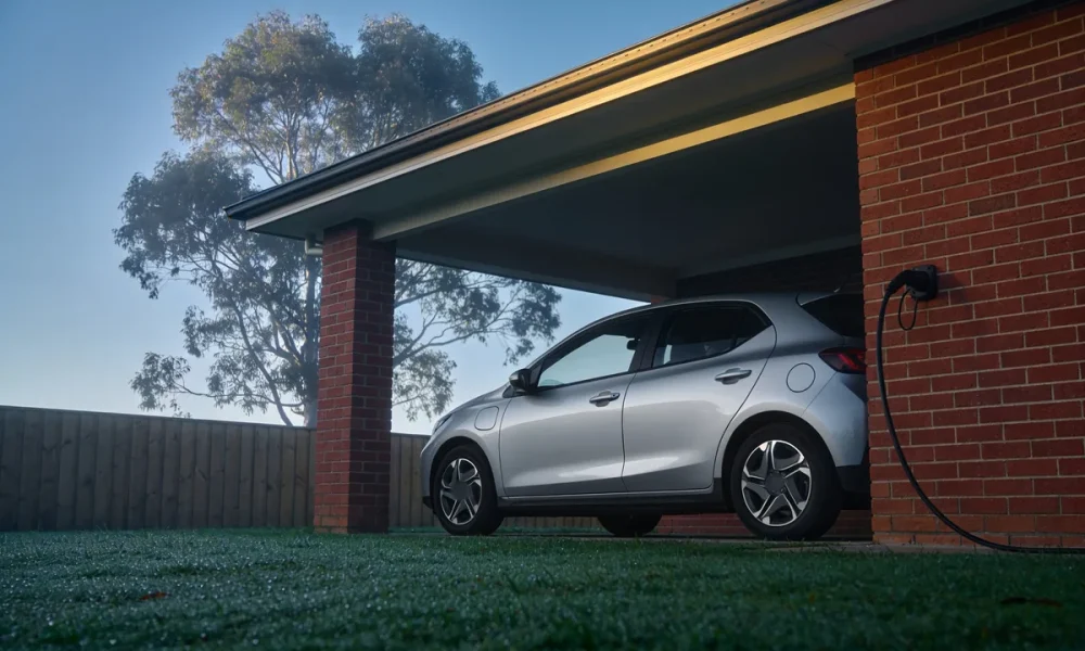 A silver compact electric hatchback parked under the carport of a red brick house on a misty morning, with a charging cable hanging from a wall socket, dew on the lawn and a gum tree behind the fence