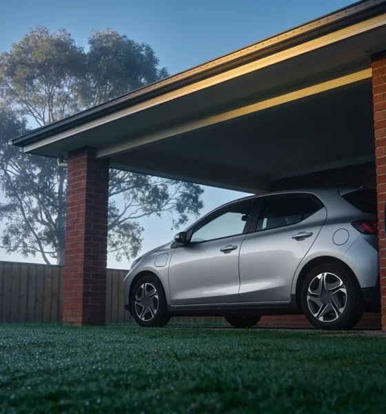 A silver compact electric hatchback parked under the carport of a red brick house on a misty morning, with a charging cable hanging from a wall socket, dew on the lawn and a gum tree behind the fence