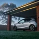 A silver compact electric hatchback parked under the carport of a red brick house on a misty morning, with a charging cable hanging from a wall socket, dew on the lawn and a gum tree behind the fence
