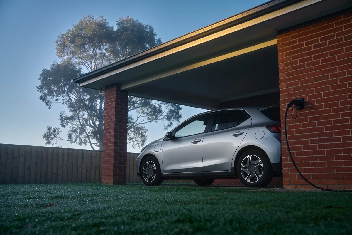 A silver compact electric hatchback parked under the carport of a red brick house on a misty morning, with a charging cable hanging from a wall socket, dew on the lawn and a gum tree behind the fence