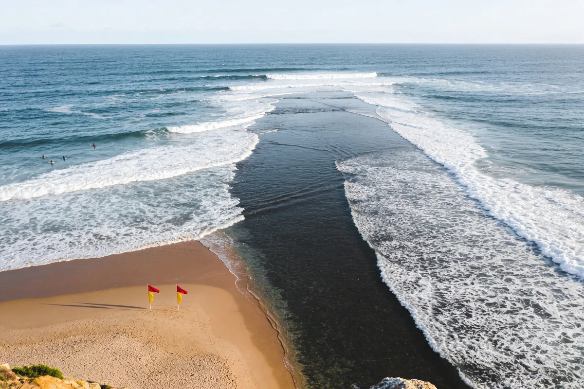 An elevated view of a surf beach with white breaking waves on both sides of a dark, flat channel of water running straight out to sea, swimmers well to one side and red and yellow flags on the sand