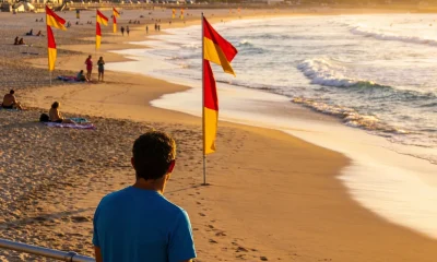 A man in a blue shirt watching the surf from a beach railing in early morning light, with red and yellow patrol flags on the sand and swimmers between them