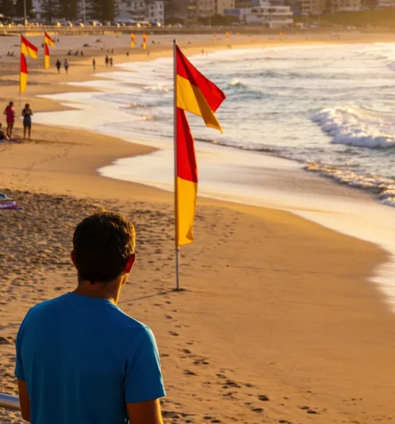 A man in a blue shirt watching the surf from a beach railing in early morning light, with red and yellow patrol flags on the sand and swimmers between them