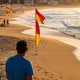 A man in a blue shirt watching the surf from a beach railing in early morning light, with red and yellow patrol flags on the sand and swimmers between them